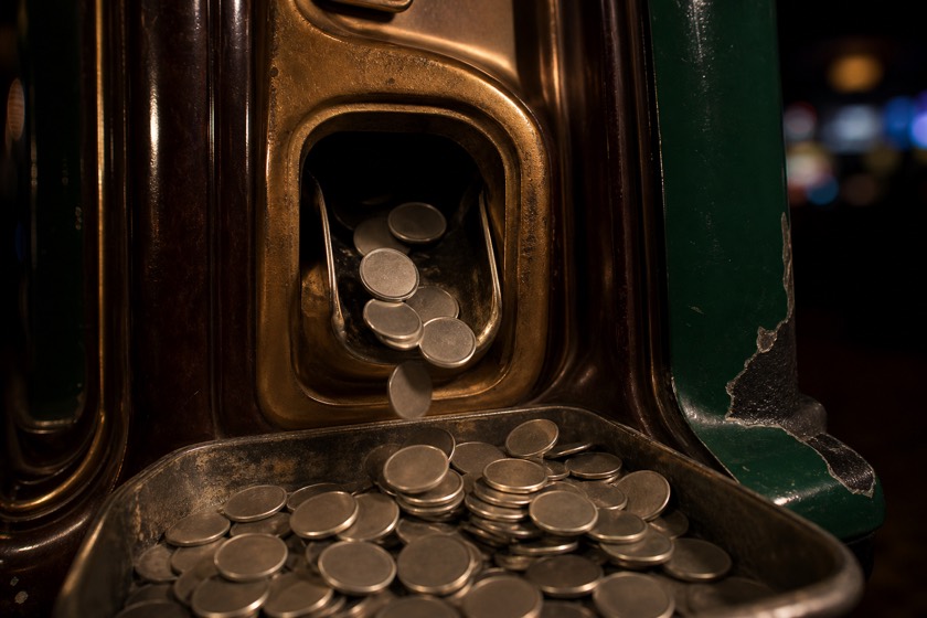 An old slot machine paying out: a cast brass chute in a bakelite cabinet with chipped green enamel flanks, blank coins tumbling into the tray below.